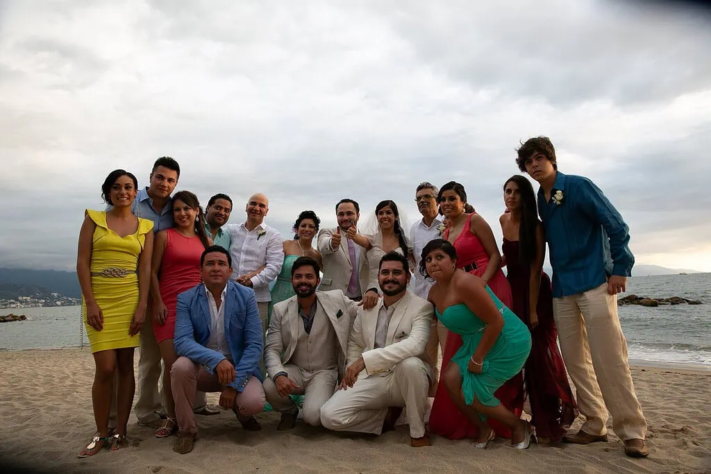 Grupo de personas en la playa durante evento familiar, con vestimenta formal y casual, en un día nublado, disfrutando de un momento de alegría y convivencia en la arena.